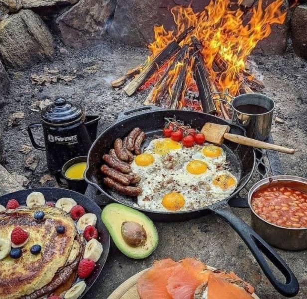 Picnic by the fire in the mountains with scrambled eggs, sausages, avocado, salmon, and pancakes with raspberries and banana.