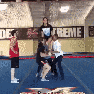 A group of guys are training. Three of them throw the girl up and she makes several turns around her axis. The guy in the white shirt falls on his back and does a backward roll.