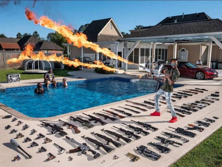 Three men by a swimming pool and a guy with many guns lying around fires from a liquid fire gun.