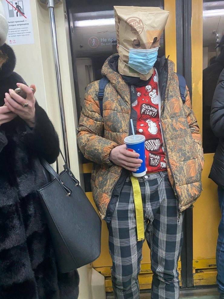 A person in a metro wearing a face mask and a paper bag on the face.