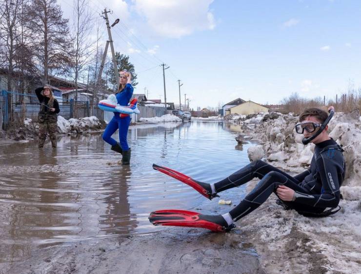 Three people standing in the street-wide deep puddle dressed like a fisher, scuba diver, and a super-woman.